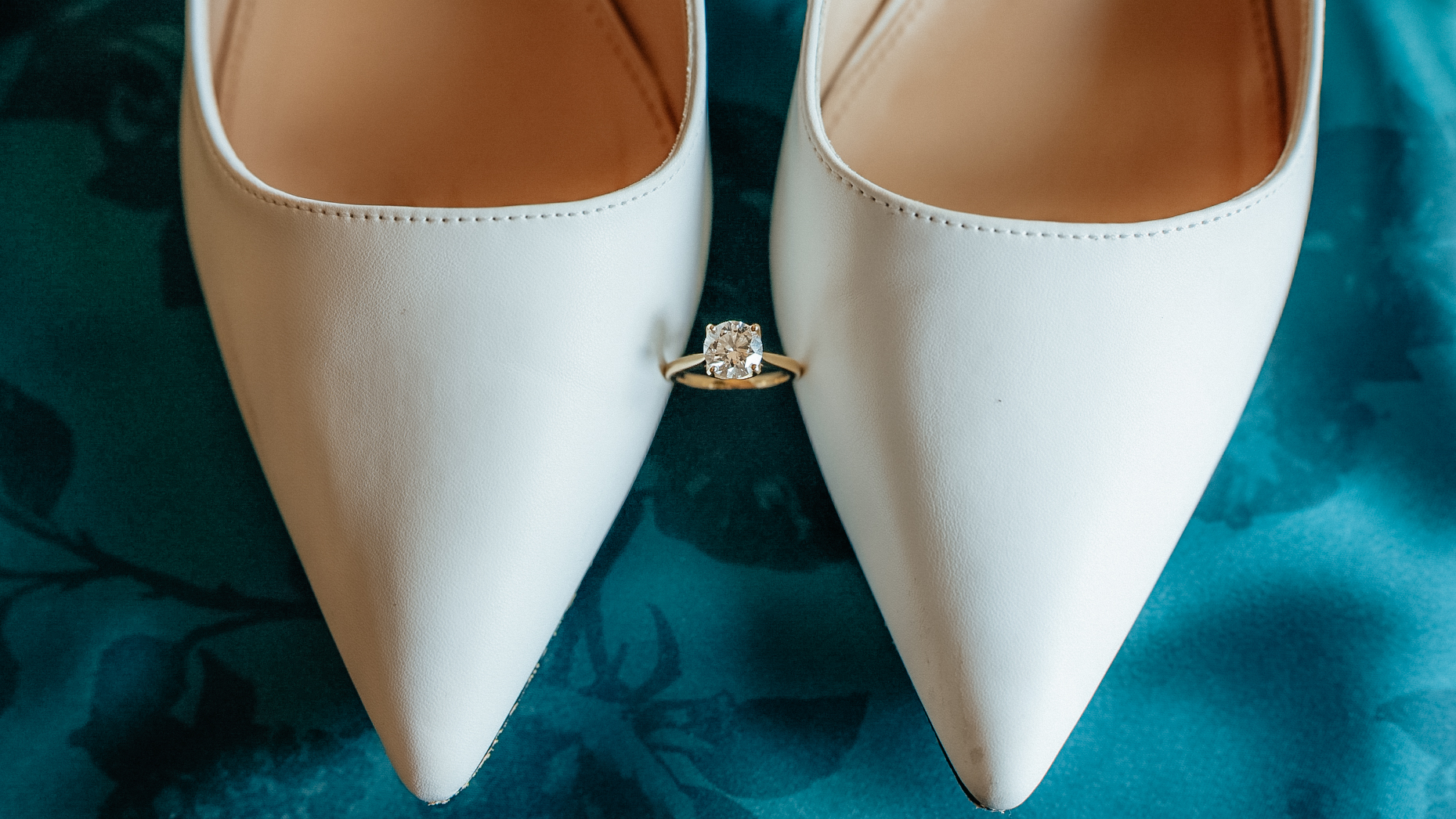 An engagement ring balanced between white bridal shoes on an emerald green background.