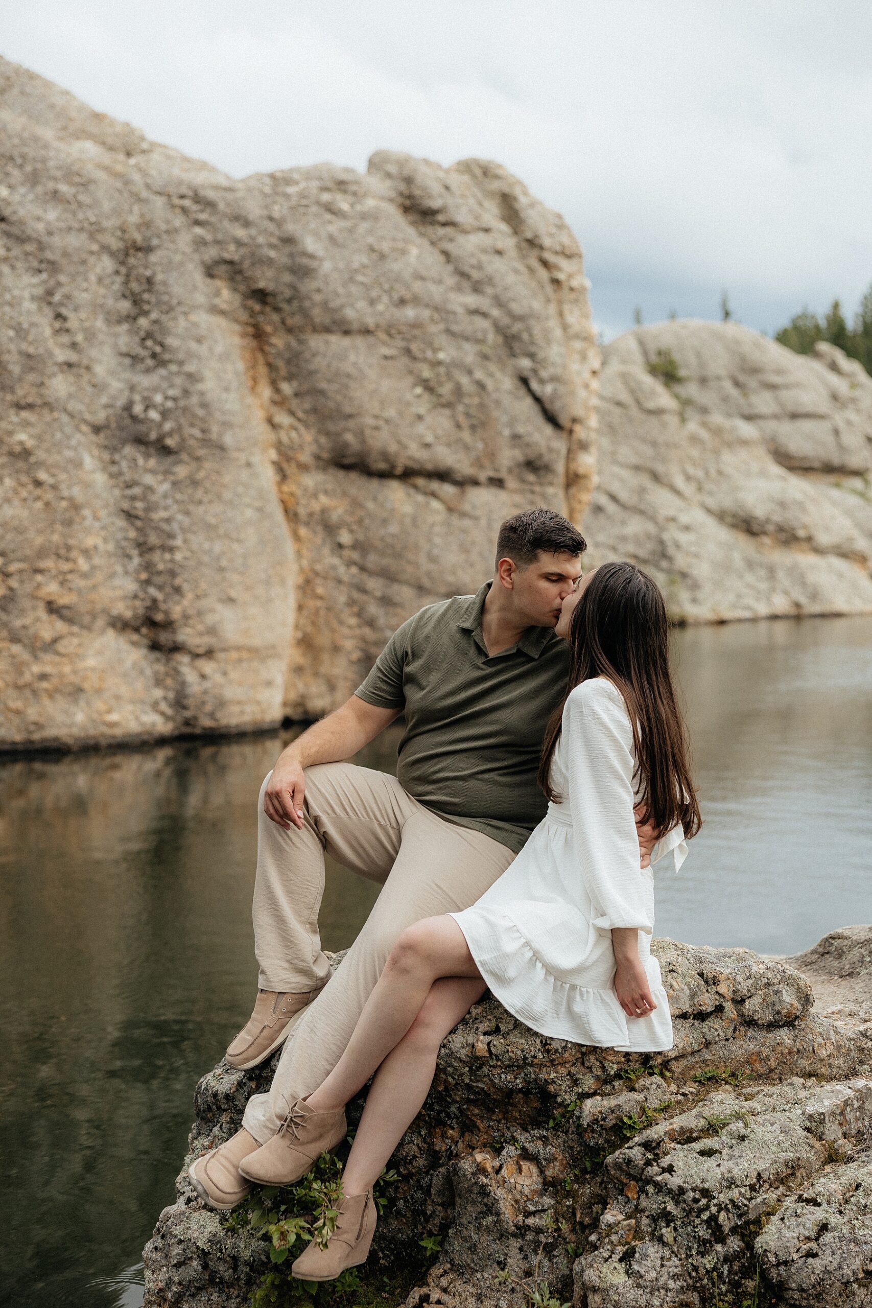 Emily + Harrison kiss while sitting on a rock with the lake behind them.