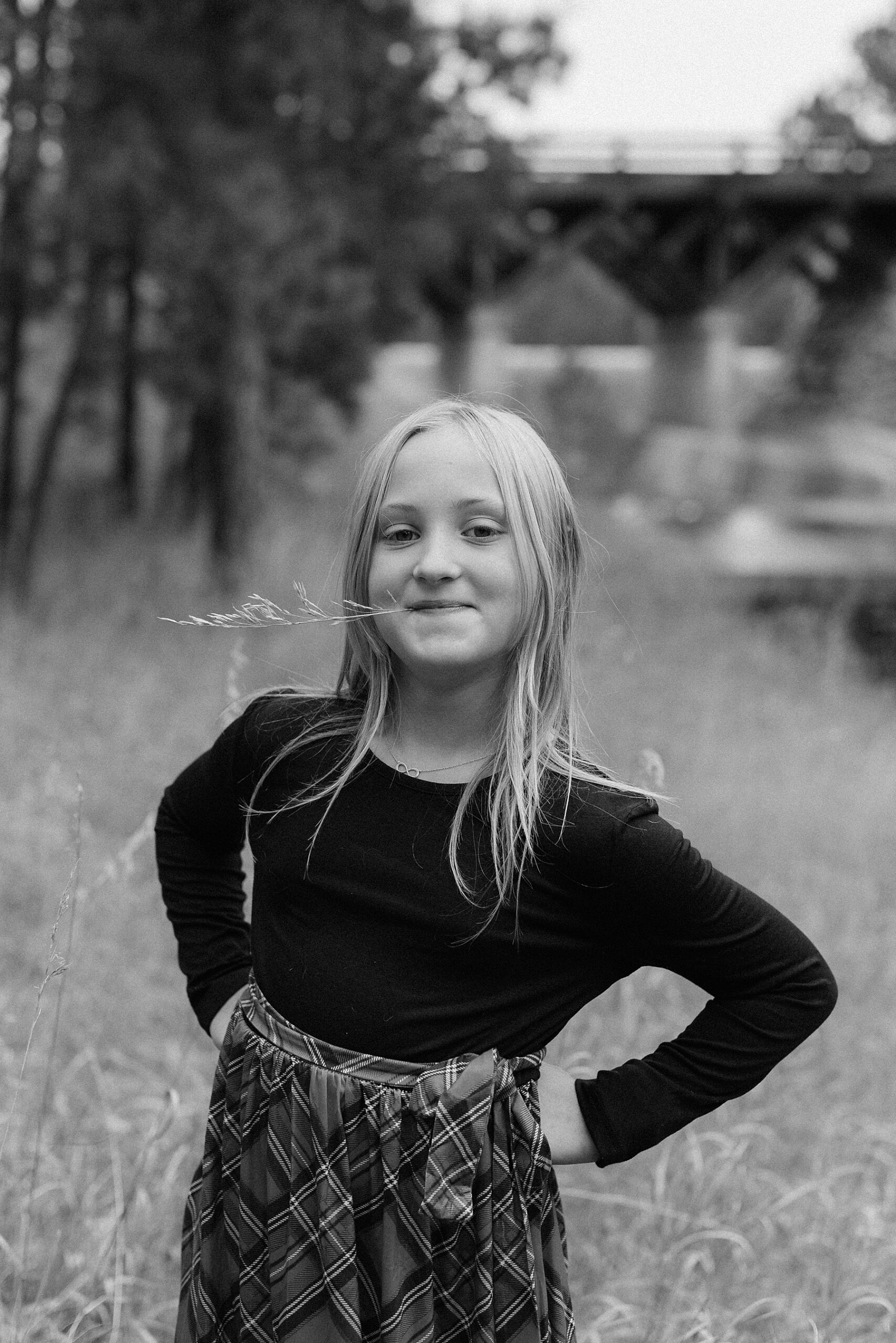 A girl standing with her hands on her hips and a piece of grass in her mouth.