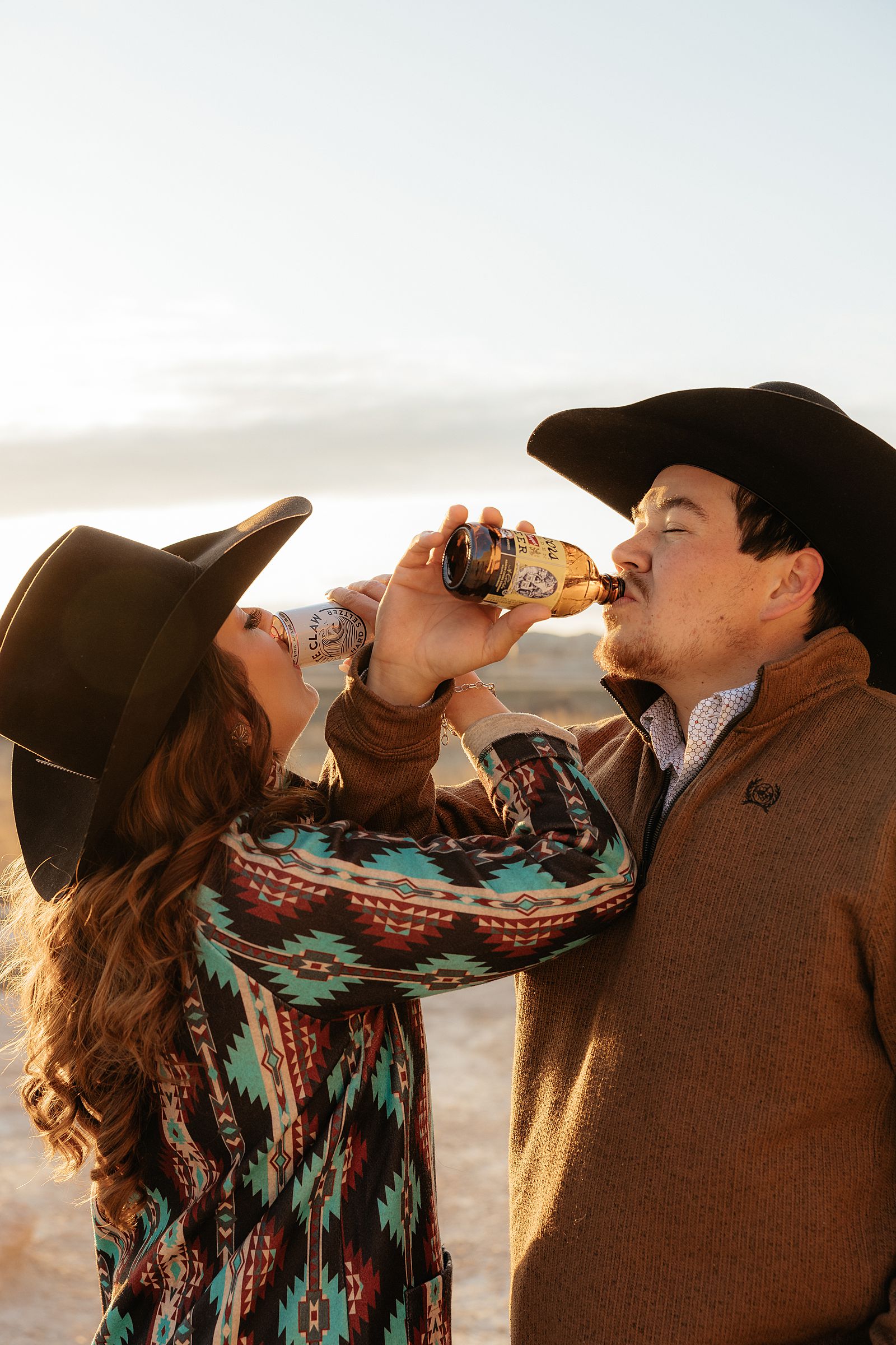 A couple wearing cowboy hats drinking beer during their engagement session.